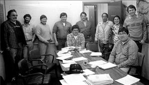 A group of men and women, mostly Native American, stand and sit around a conference table covered in stacks of paper. 